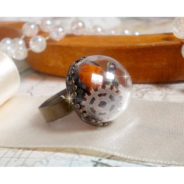 Bague Majest&eacute; Steampunk aux rouages m&eacute;caniques &agrave; laquelle s&rsquo;ajoute une cl&eacute; et un strass de couleur Siam et un strass en verre Dor&eacute;. Le tout est mis sous un globe en verre sur une bague r&eacute;glable laiton