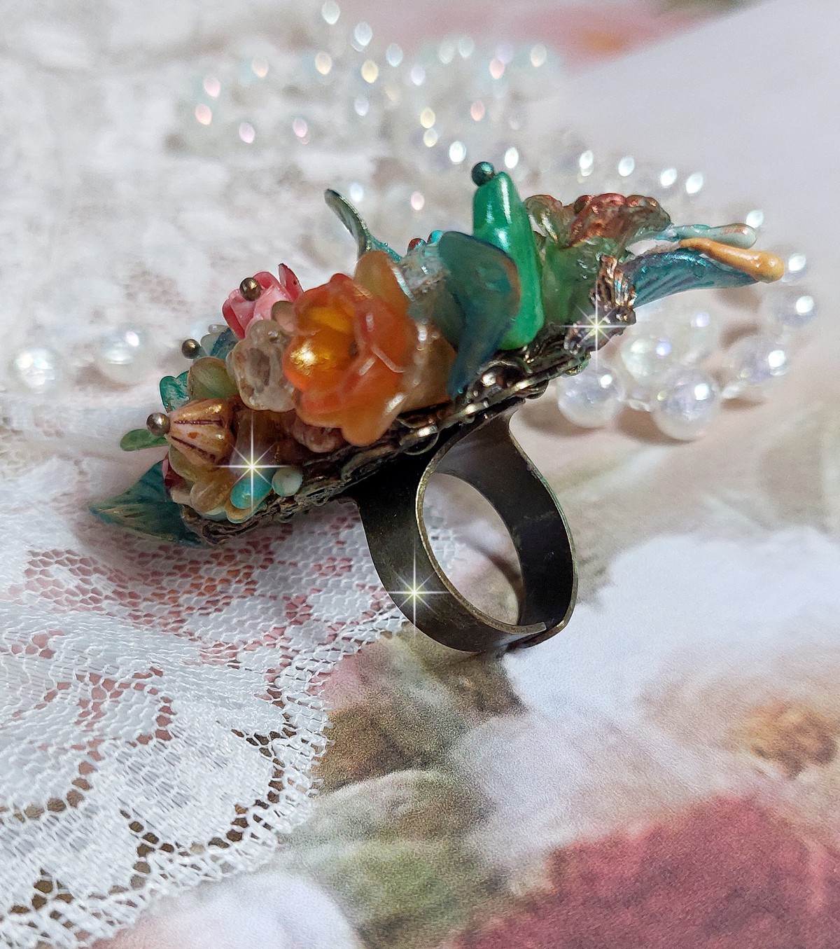 Bague Patiflo d’une composition florale créée avec des cristaux, fleurs en Résine, perles, clochettes, pensées, boutons de rose en Verre, estampes et coupelles. Le tout est monté sur une bague estampe