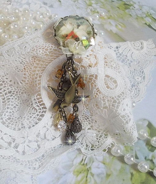 Broche Colombes aux plumages blancs avec des breloques filigran&eacute;es gouttes et lisses, perles rondes et n&oelig;ud en cristal, ruban organza cr&egrave;me, pendentif plaqu&eacute; or avec un zircon sur une broche en m&eacute;tal