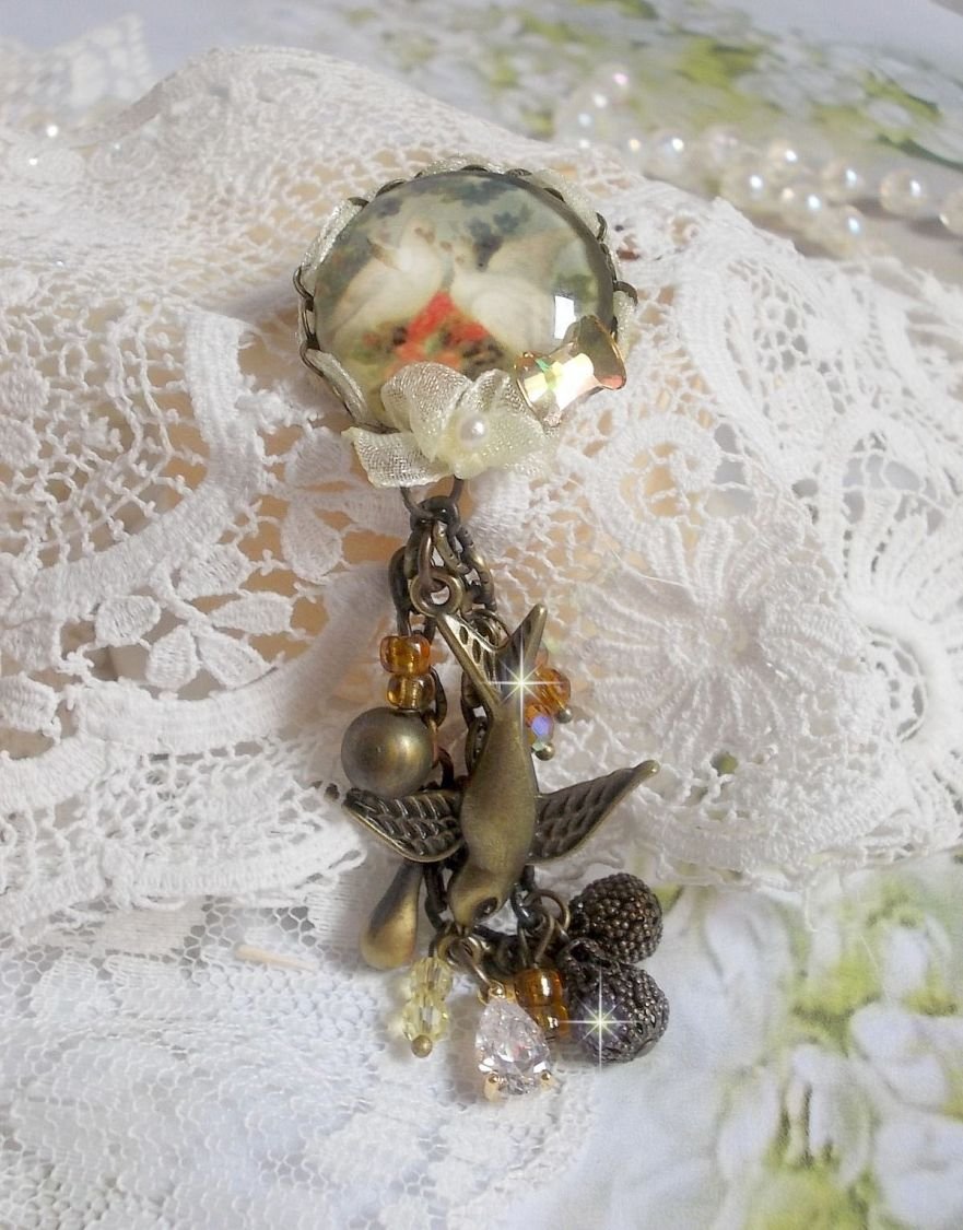 Broche Colombes aux plumages blancs avec des breloques filigranées gouttes et lisses, perles rondes et nœud en cristal, ruban organza crème, pendentif plaqué or avec un zircon sur une broche en métal