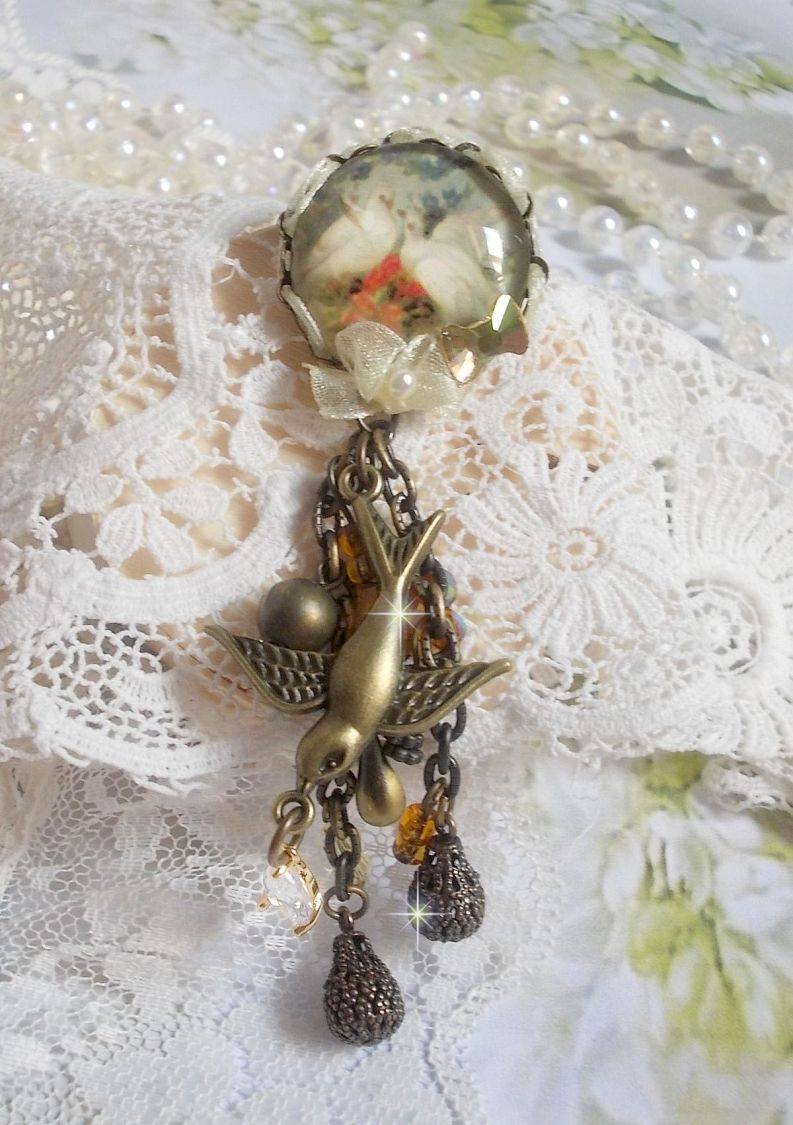 Broche Colombes aux plumages blancs avec des breloques filigranées gouttes et lisses, perles rondes et nœud en cristal, ruban organza crème, pendentif plaqué or avec un zircon sur une broche en métal