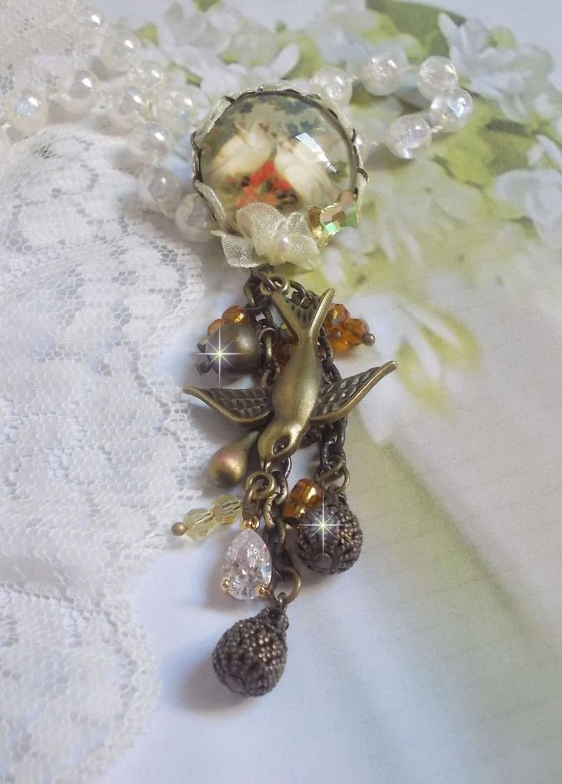 Broche Colombes aux plumages blancs avec des breloques filigranées gouttes et lisses, perles rondes et nœud en cristal, ruban organza crème, pendentif plaqué or avec un zircon sur une broche en métal