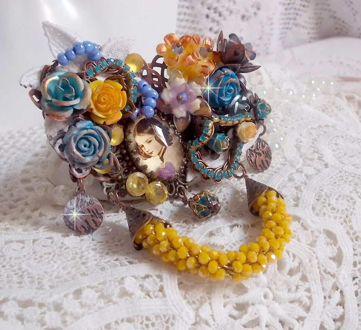 Broche Eclats Pétillants d’inspiration Baroque, ornée de fleurs en résine autour d’un cabochon strassés et perlés sur estampes, cristaux, breloques, perle cloisonnée, perles jaunes en verre et roses. 