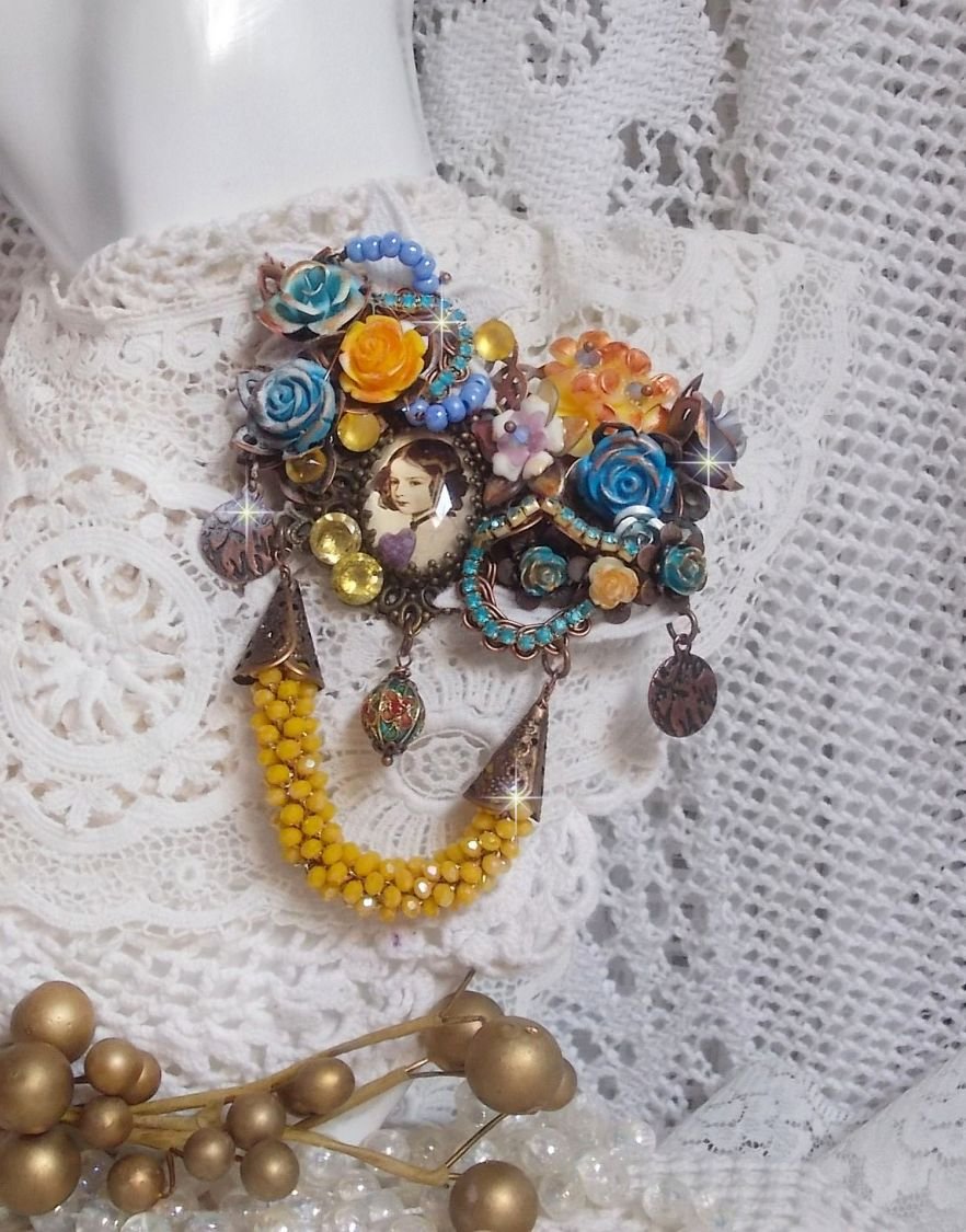 Broche Eclats Pétillants d’inspiration Baroque, ornée de fleurs en résine autour d’un cabochon strassés et perlés sur estampes, cristaux, breloques, perle cloisonnée, perles jaunes en verre et roses. 